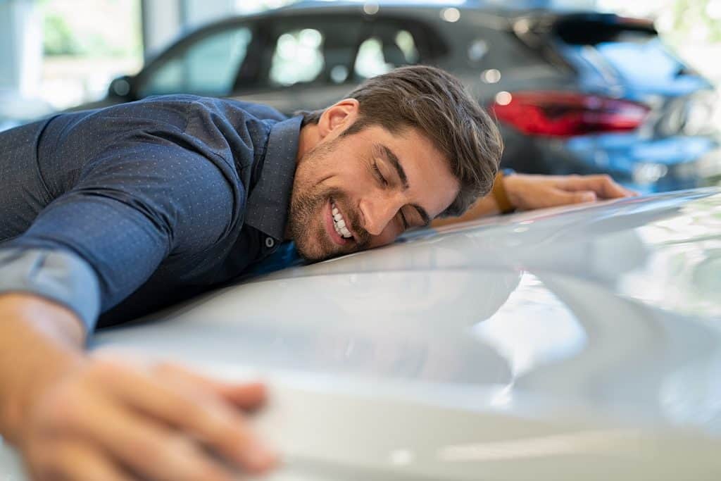 Happy young man hugging his new car in showroom. 