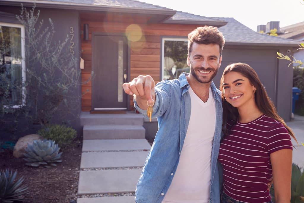 Portrait Of Couple Standing Outdoors In Front Of House With For Sale Sign In Garden Holding Keys